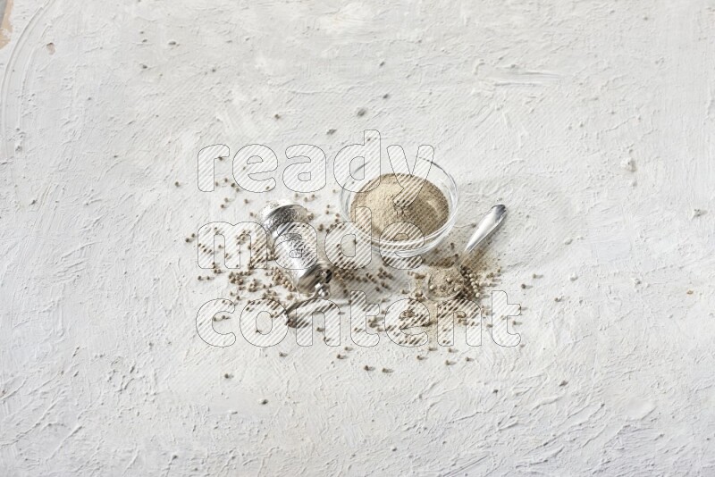 A glass bowl full of white pepper powder with white pepper beads and a metal grinder on textured white flooring