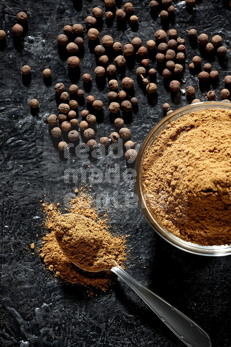 A glass bowl and metal spoon full of allspice powder and whole balls spreaded on a black flooring