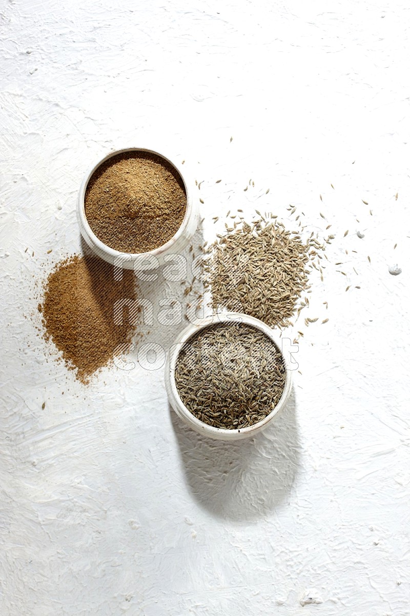 2 beige bowls full of cumin seeds and powder with spilled powder and seeds on textured white flooring