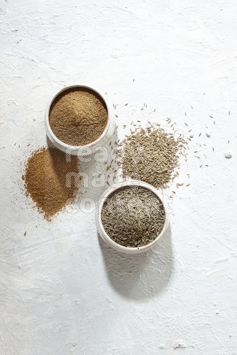 2 beige bowls full of cumin seeds and powder with spilled powder and seeds on textured white flooring