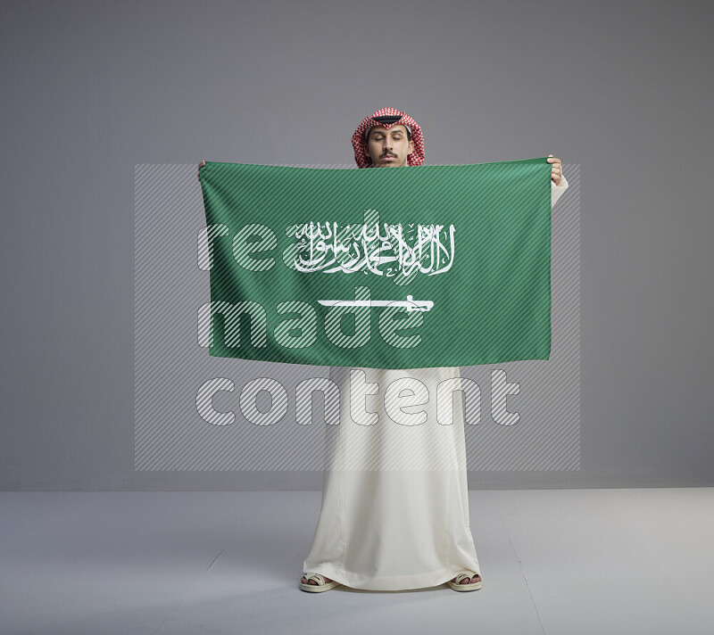 A saudi man standing wearing thob and red shomag holding big saudi flag on gray background