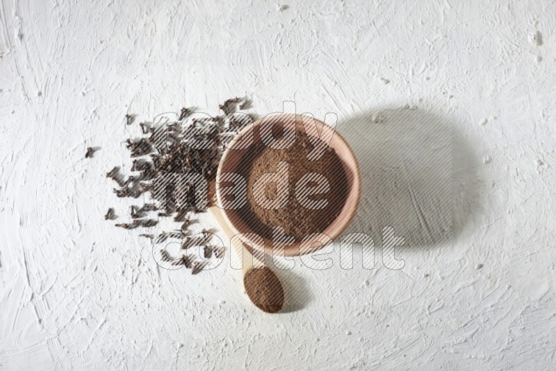 A wooden bowl and wooden spoon full of cloves powder with cloves spread on textured white flooring