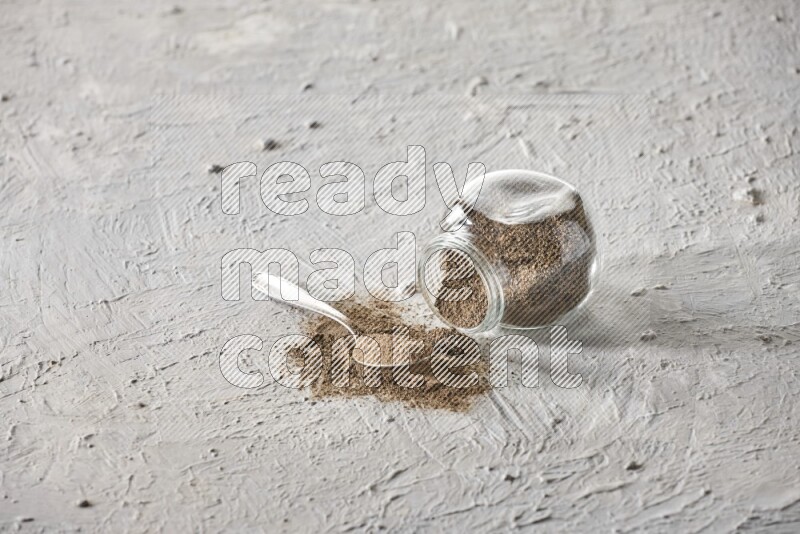 Flipped glass spice jar full of black pepper powder with a metal spoon full of it on textured white flooring