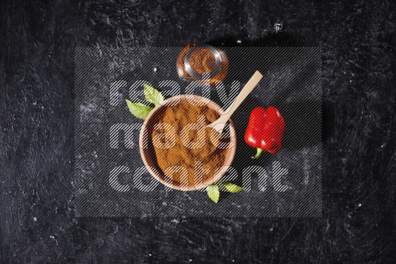 A wooden bowl full of ground paprika powder with a glass jar beside it and a red bell pepper on black background