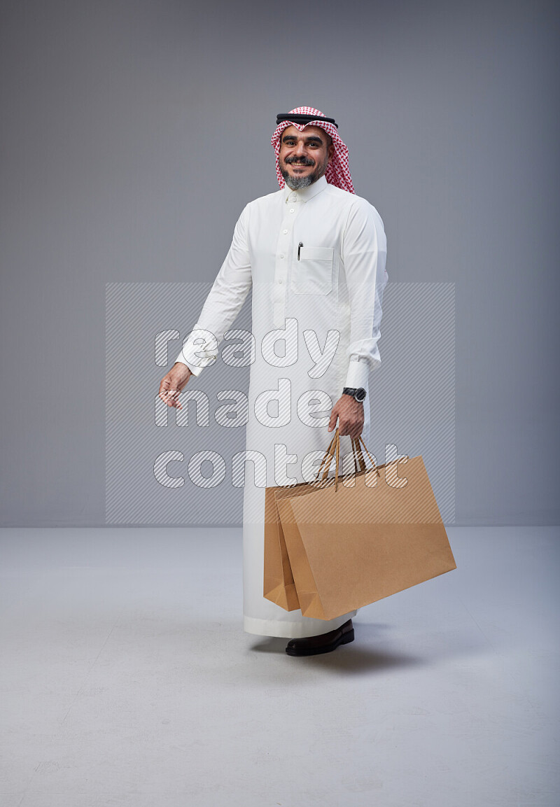 Saudi man Wearing Thob and red Shomag standing holding shopping bag on Gray background