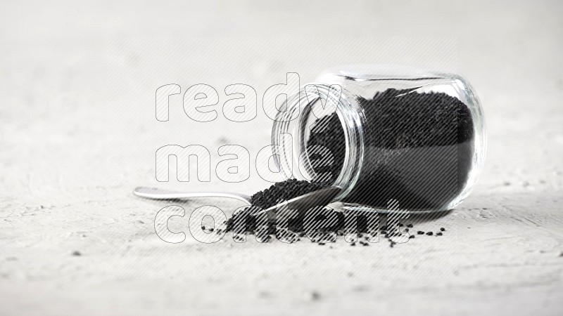 A glass spice jar and a metal spoon full of black seeds and the jar flipped and seeds spread on a textured white flooring