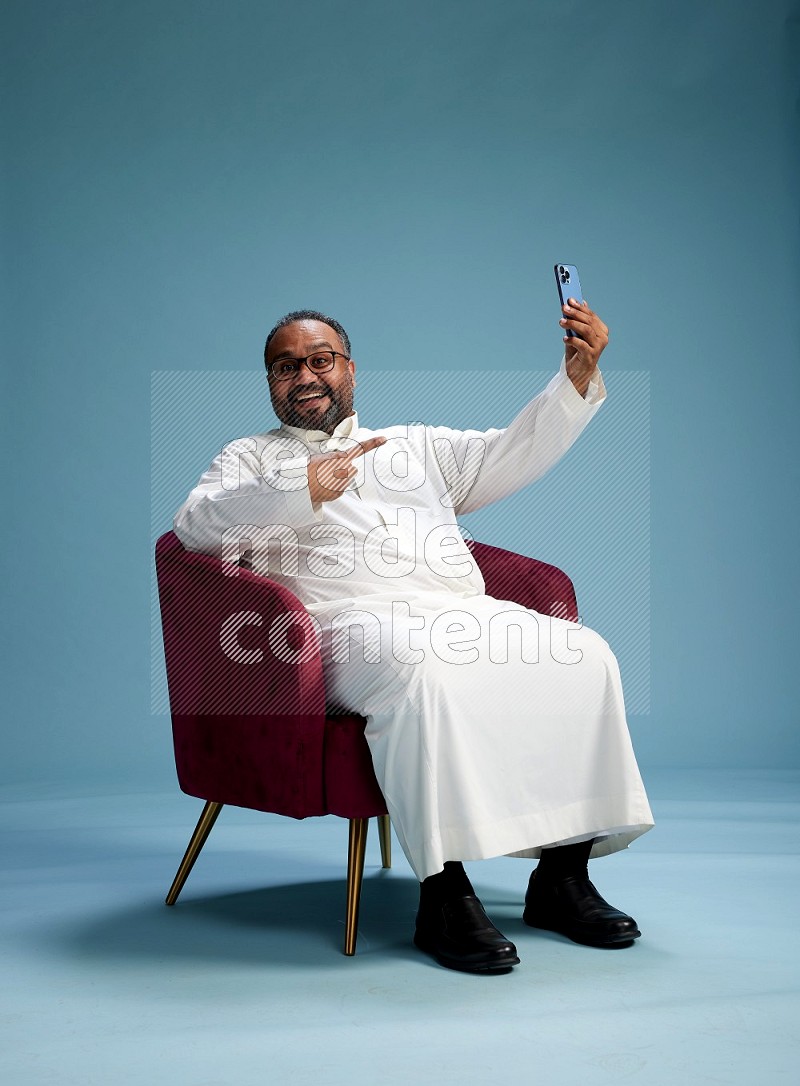 Saudi Man without shimag sitting on chair taking selfie on blue background