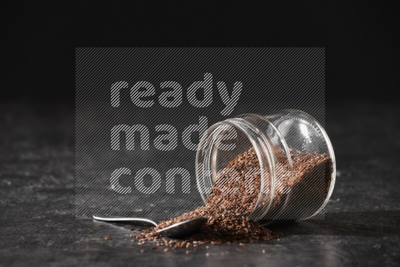 A glass jar full of flaxseeds flipped and seeds spread out with a metal spoon full of the seeds on a textured black flooring
