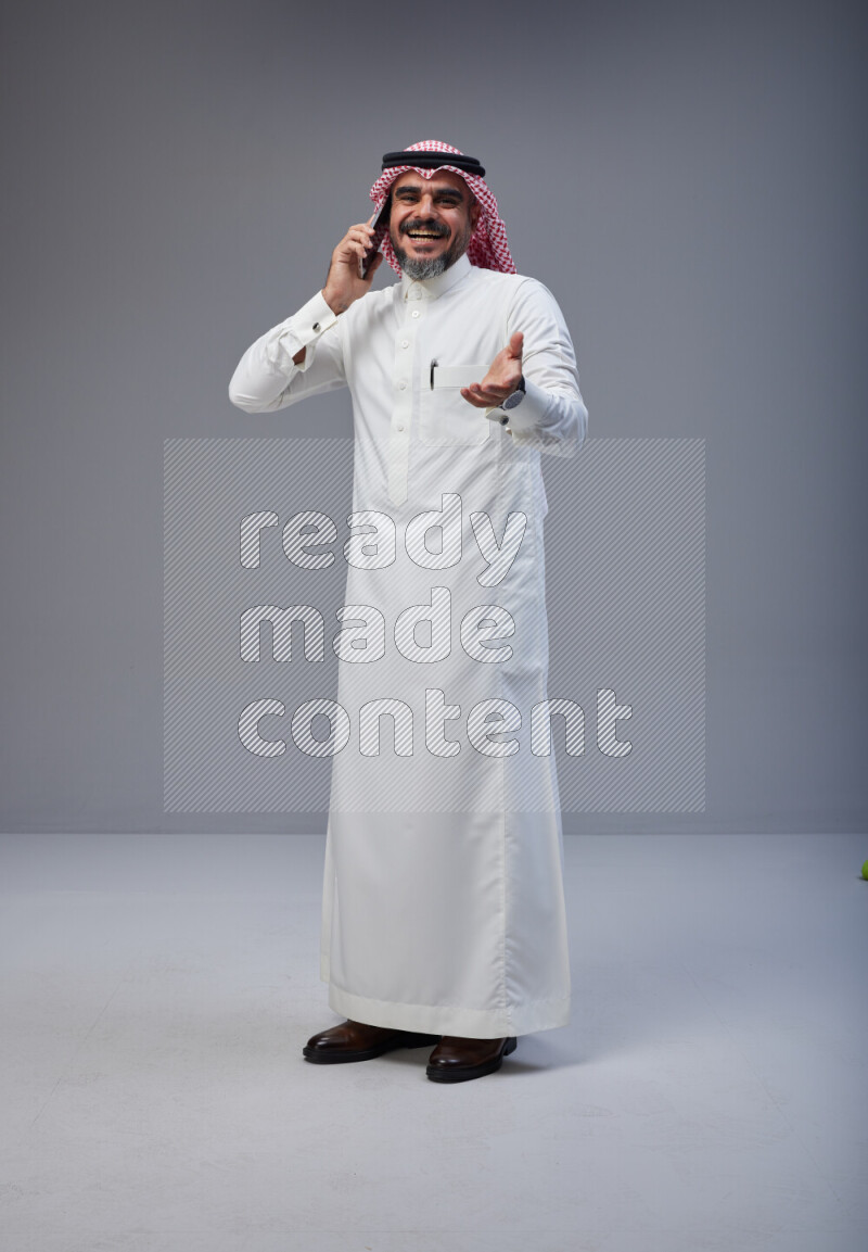 Saudi man Wearing Thob and red Shomag standing talking on phone on Gray background