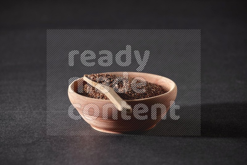 A wooden bowl full of flaxseeds with wooden spoon full of the seeds on it on a black flooring