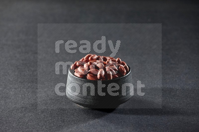 A black pottery bowl full of red skin peanuts on a black background in different angles