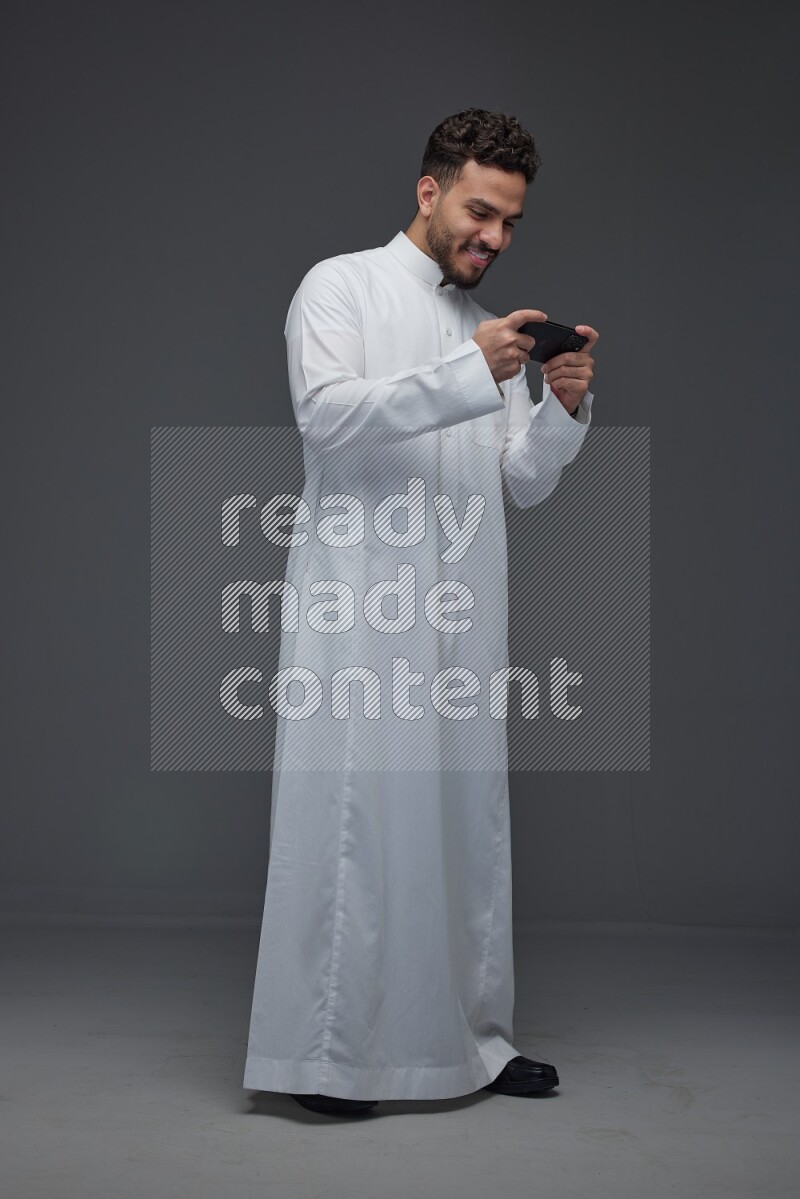 A Saudi man wearing Thobe and playing using his phone horizontally playing video games while standing and making different poses eye level on a gray background