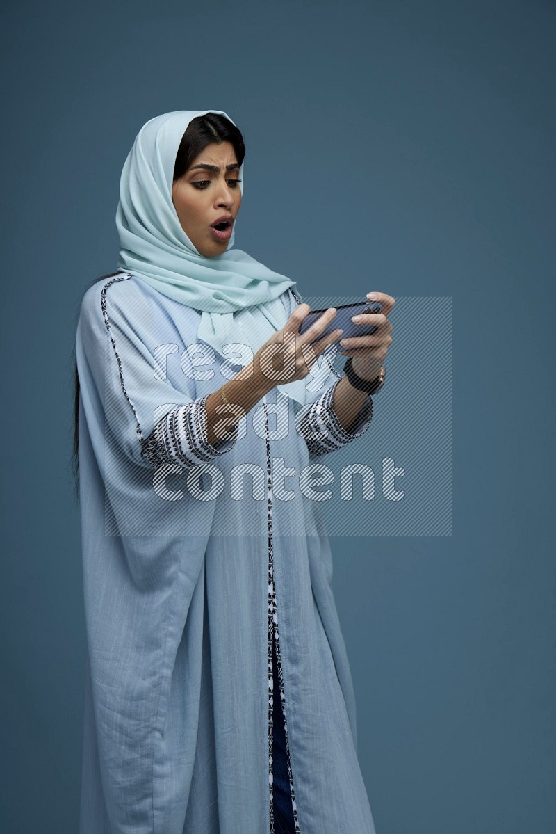A Saudi woman Playing a Game on her phone on a blue background wearing a blue Abaya with hijab