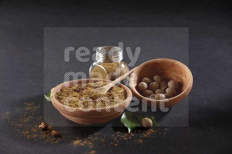 A wooden bowl with a spoon in it and glass spice jar all full of nutmeg powder and a wooden bowl full of whole nutmeg seeds on a black flooring