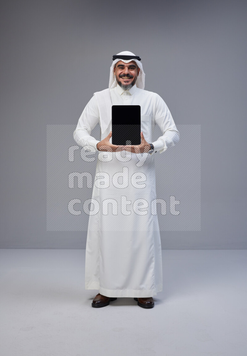 Saudi man Wearing Thob and white Shomag standing showing tablet to camera on Gray background