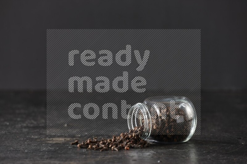 A flipped glass spice jar full of cloves and cloves came out of it on textured black flooring