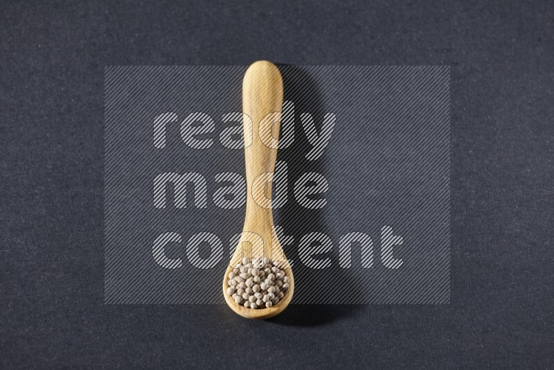 A wooden spoon full of white pepper beads on black flooring