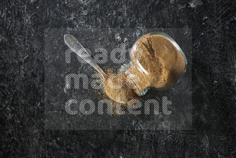A flipped glass spice jar and a metal spoon full of cumin powder and powder spilled out on a textured black flooring