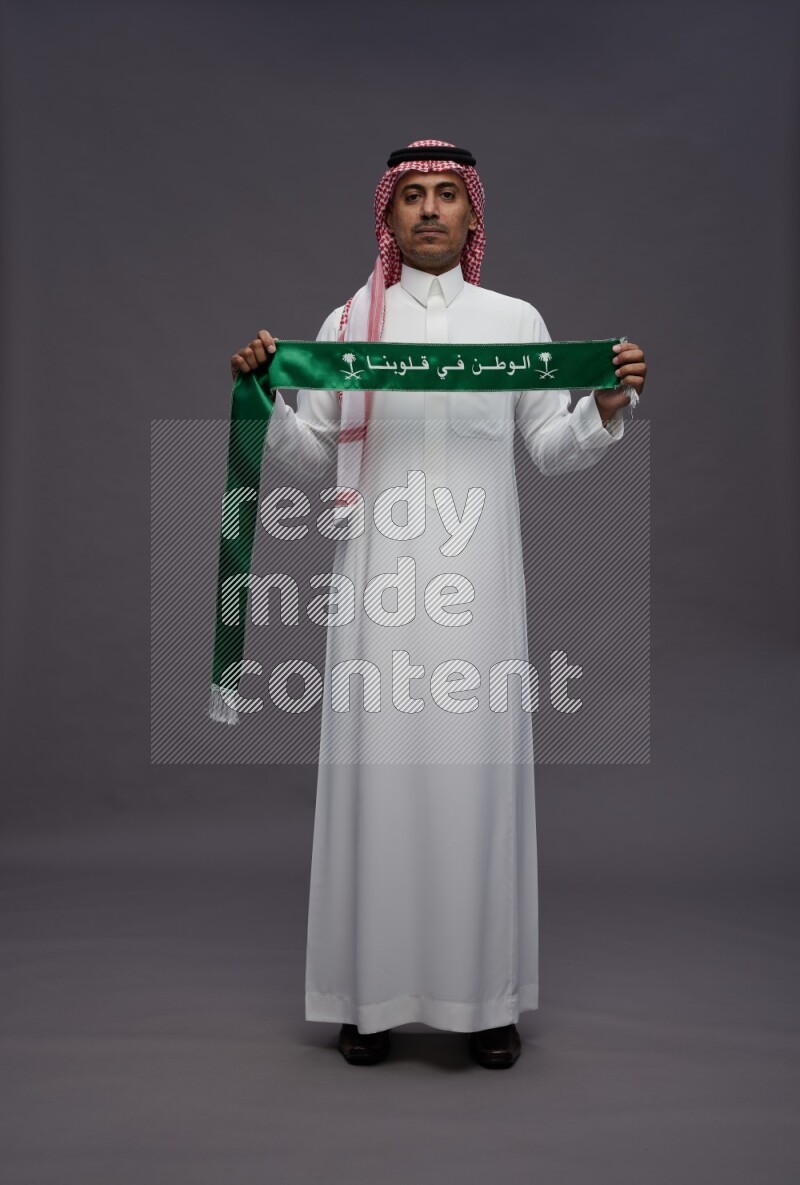 Saudi man wearing thob and shomag standing holding Saudi flag on gray background