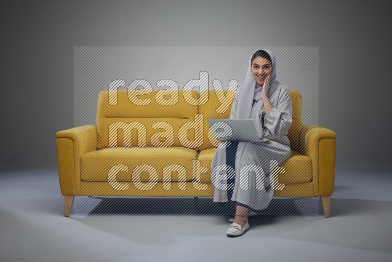 A Saudi woman wearing a light gray Abaya and head scarf sitting on a yellow sofa and using her laptop eye level on a grey background