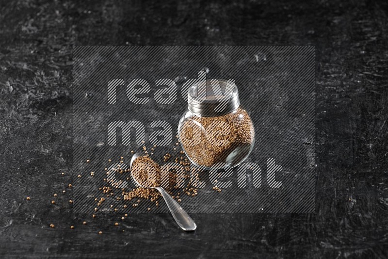 A glass spice jar and a metal spoon full of mustard seeds on a textured black flooring