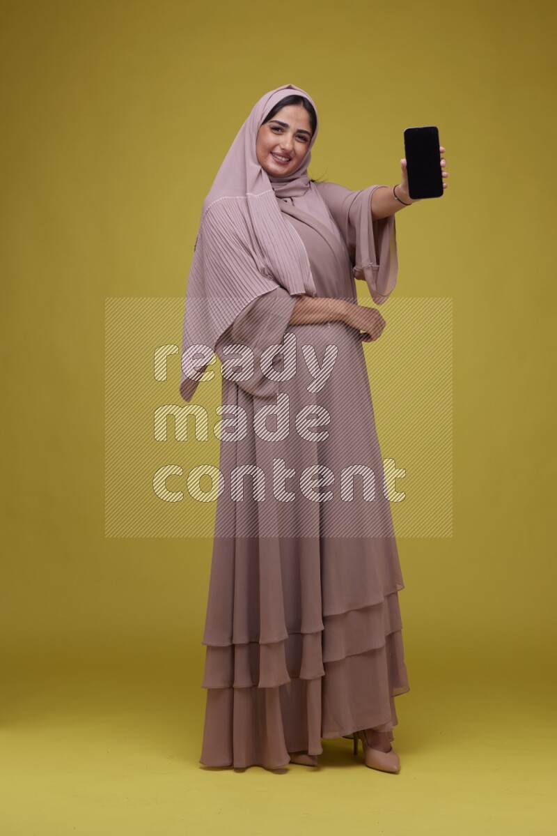 A woman Showing her Phone screen on a Yellow Background wearing Brown Abaya with Hijab
