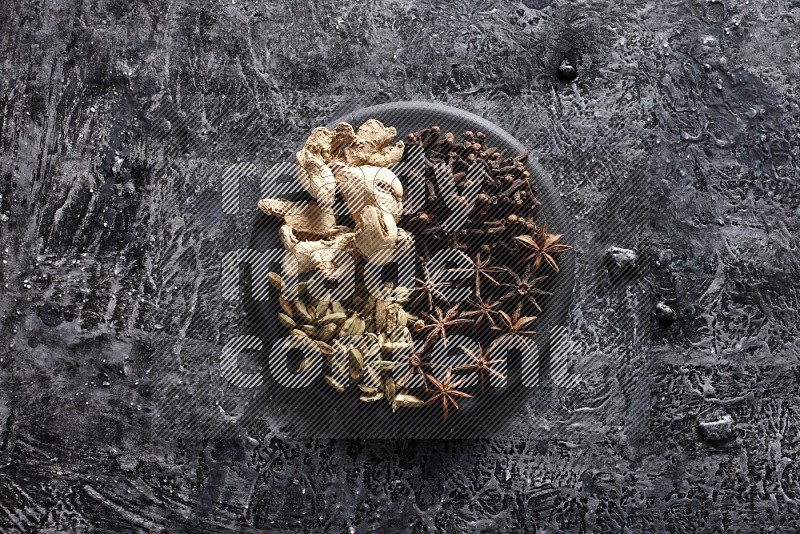 Ginger, cloves, star anise and cardamom on a black plate on textured black background