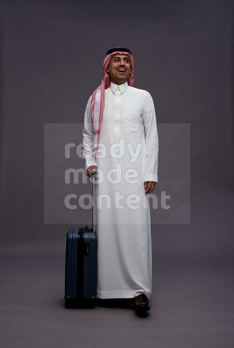 Saudi man wearing thob and shomag standing holding bag on gray background