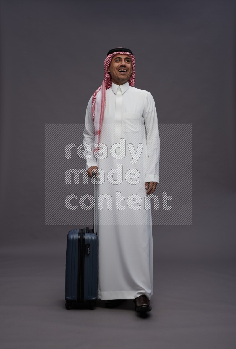 Saudi man wearing thob and shomag standing holding bag on gray background