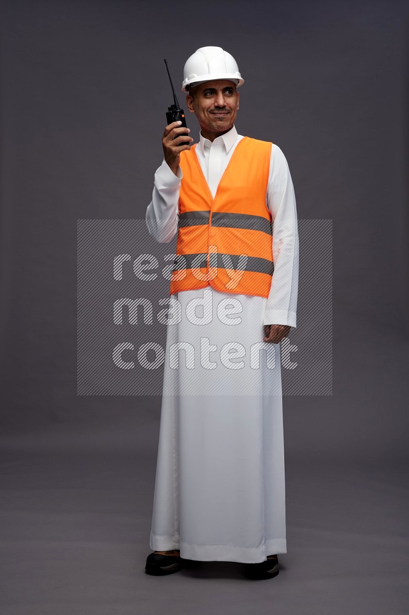 Saudi man wearing thob with engineer vest standing holding walkie-talkie on gray background