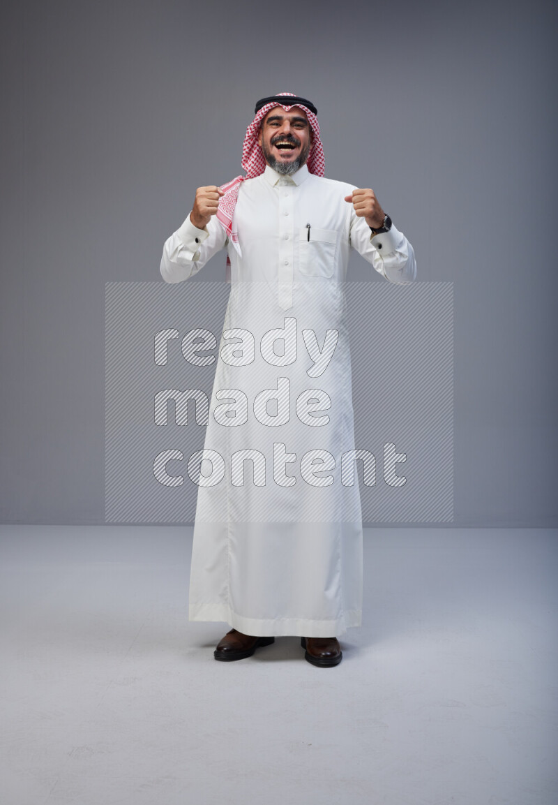 Saudi man Wearing Thob and red white Shomag standing interacting with the camera on Gray background