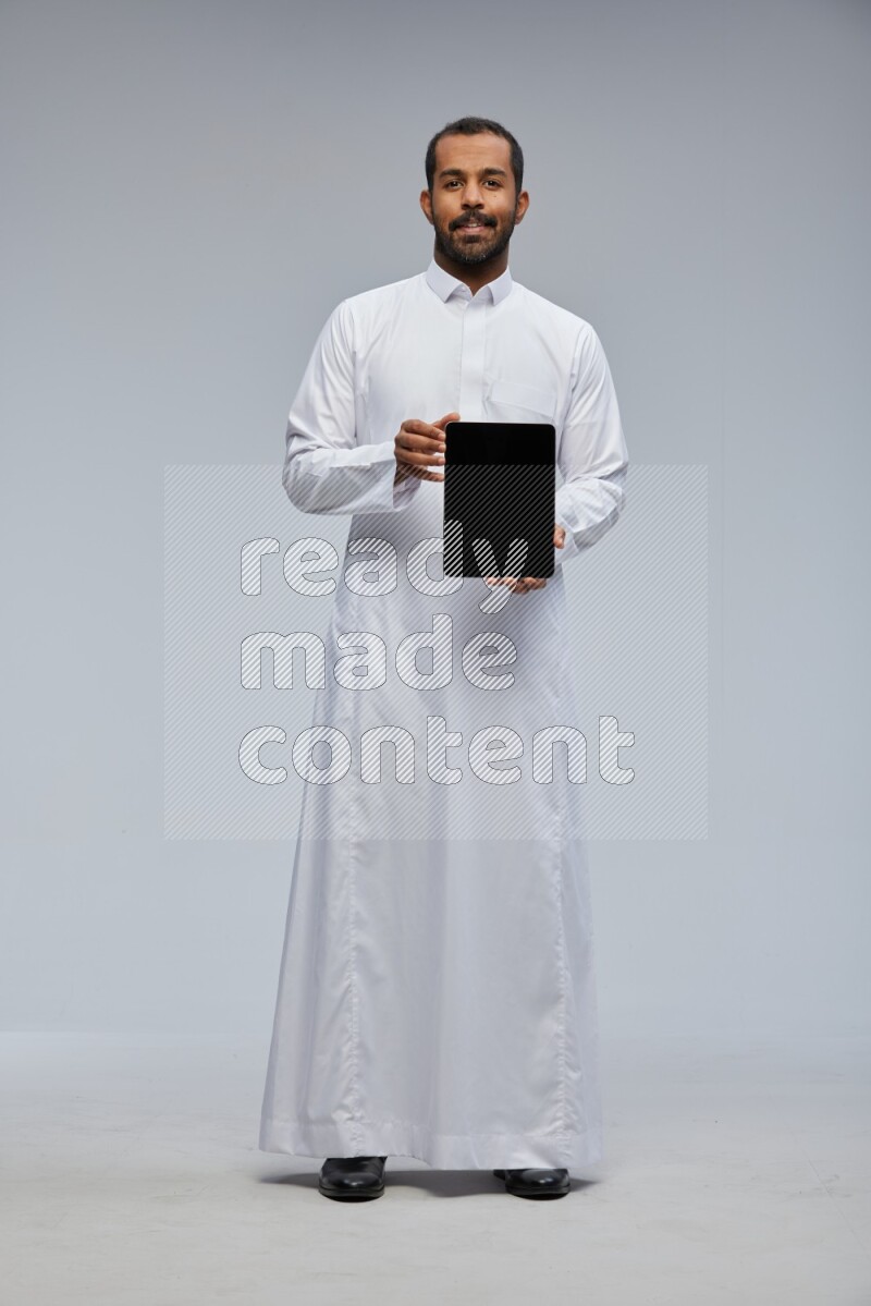 Saudi man wearing Thob standing showing tablet to camera on Gray background