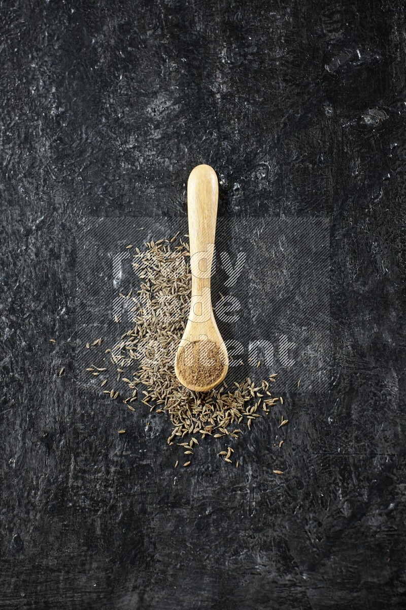 A wooden spoon full of cumin powder and cumin seeds spreaded on textured black flooring
