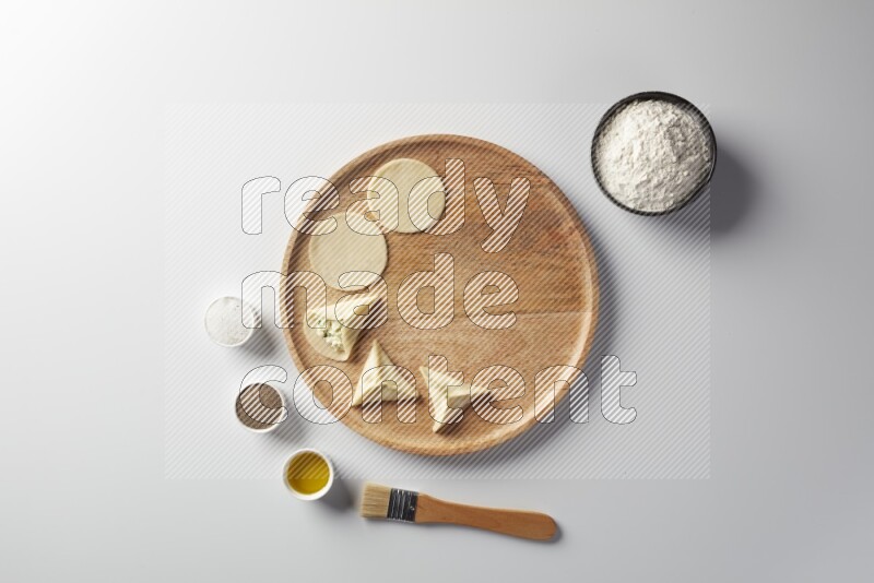 two closed sambosas and one open sambosa filled with cheese while flour, salt, black pepper and oil with oil brush aside in a wooden dish on a white background
