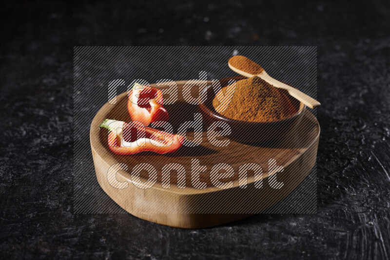 A wooden bowl full of ground paprika powder and sliced red bell pepper beside it, all on a wooden tray on black background