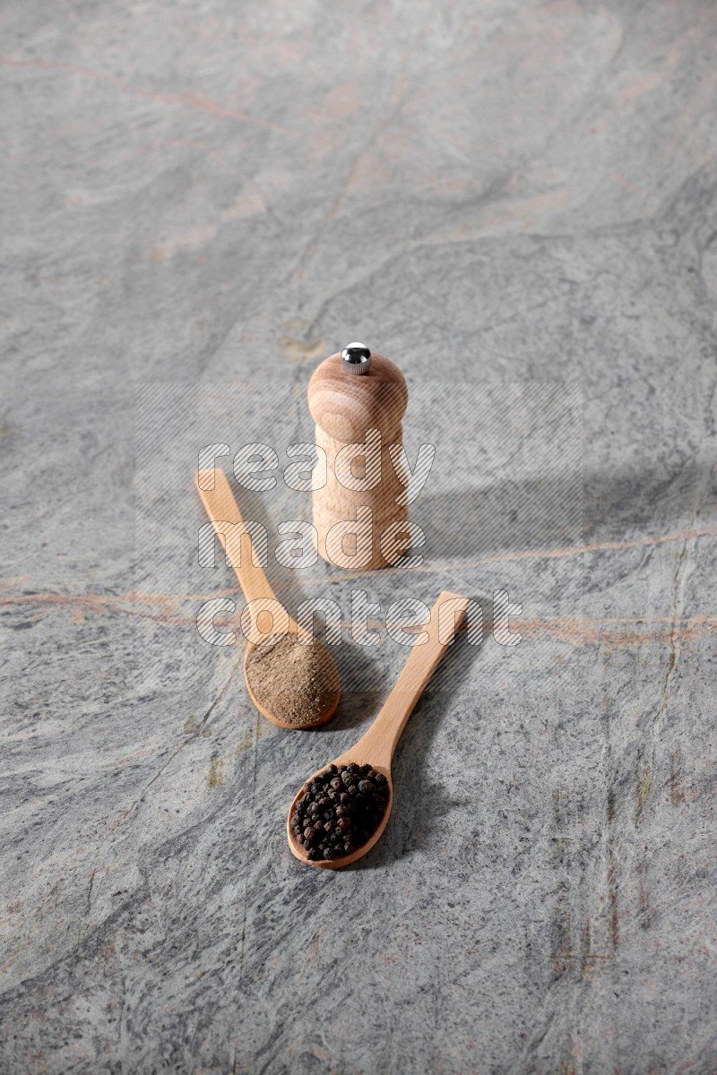2 wooden spoons, one full of black pepper powder and the other full of black pepper beads and wooden grinder on a marble flooring