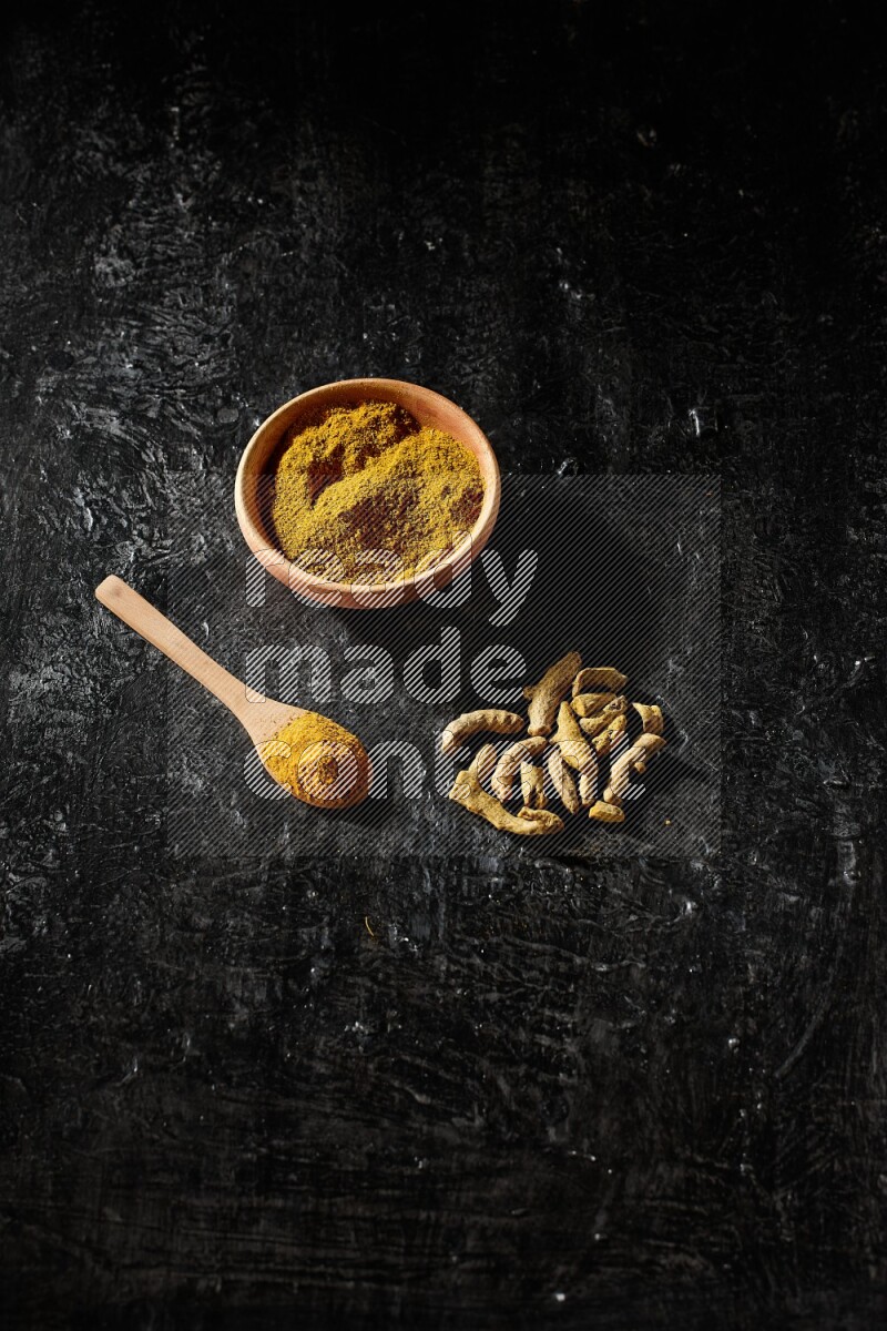 A wooden bowl and a wooden spoon full of turmeric powder with dried turmeric fingers on textured black flooring