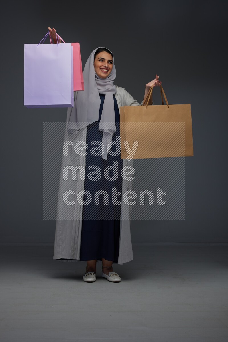 A Saudi woman wearing a light gray Abaya and head scarf standing and holding shopping bags on a grey background