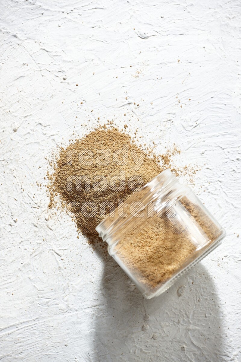 A flipped glass jar full of cumin powder and powder spilled out on textured white flooring
