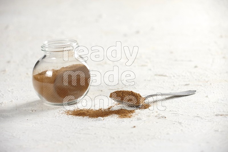 Herbs glass jar full of cinnamon powder with a metal spoon full of powder on a textured white background