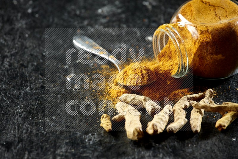 A flipped glass spice jar and metal spoon full of turmeric powder and powder spilled out of it with dried whole fingers on textured black flooring
