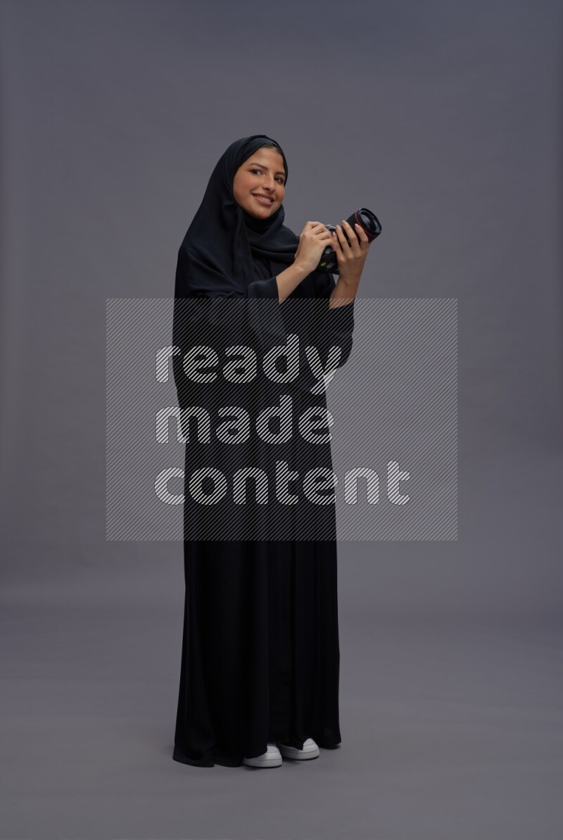 Saudi woman wearing Abaya standing holding Camera on gray background