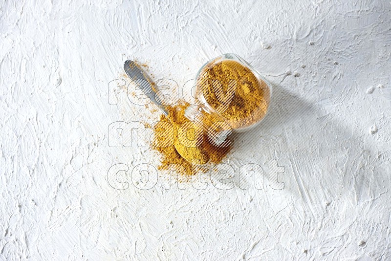 A flipped glass spice jar and a metal spoon full of turmeric powder and powder spilled out of it on textured white flooring