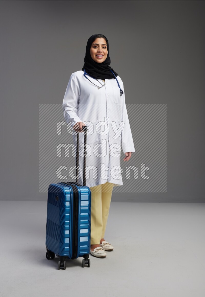 Saudi woman wearing lab coat with stethoscope standing holding Travel bag on Gray background