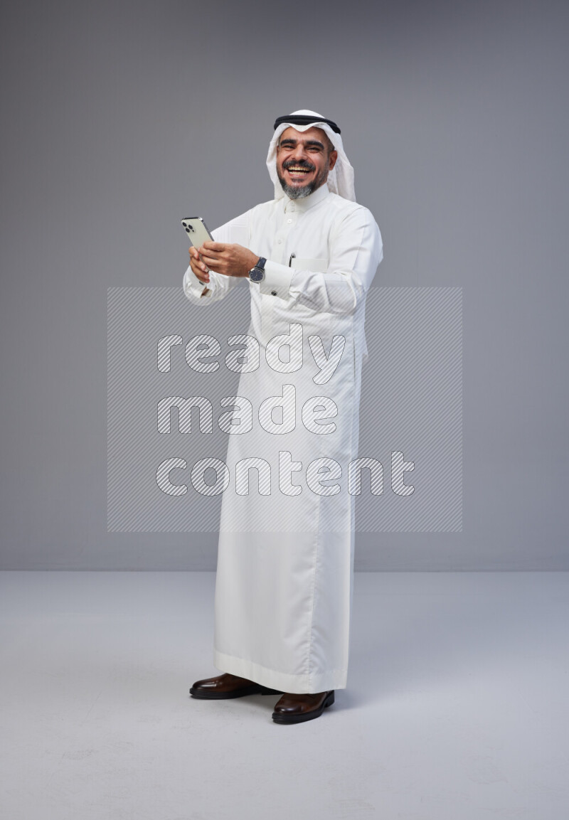 Saudi man Wearing Thob and white Shomag standing texting on phone on Gray background