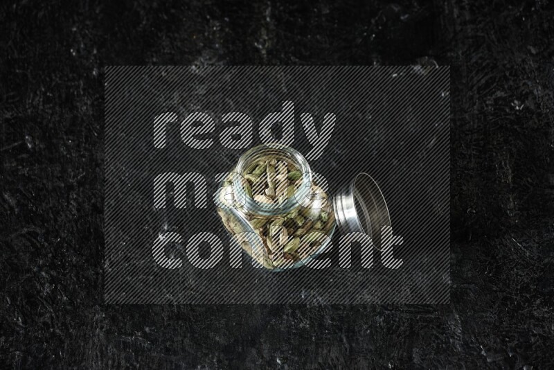 A glass spice jar full of cardamom seeds on textured black flooring