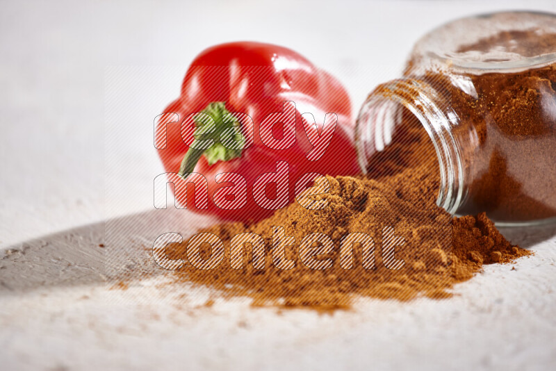 A glass jar full of ground paprika powder flipped with some spilling powder on white background