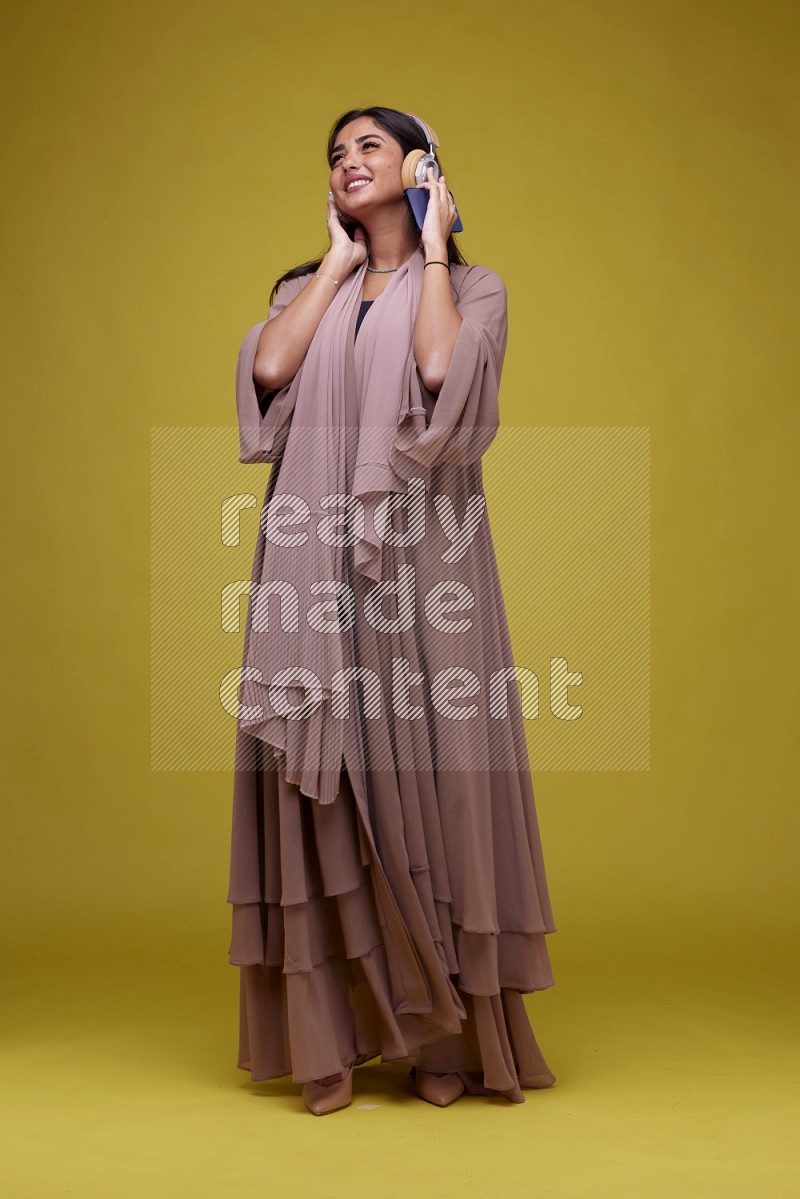 A Saudi woman Listing to Music on a Yellow Background wearing Brown Abaya