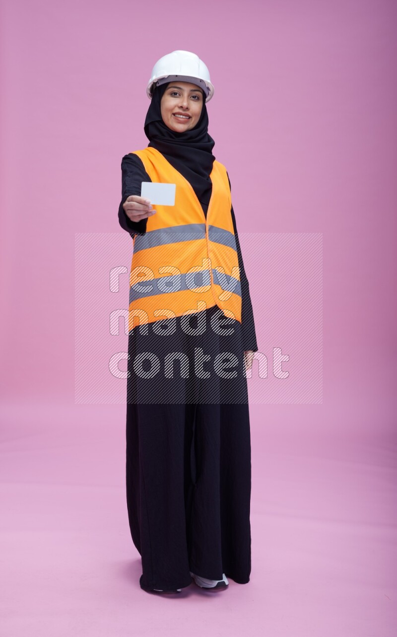 Saudi woman wearing Abaya with engineer vest and helmet standing holding ATM card on pink background