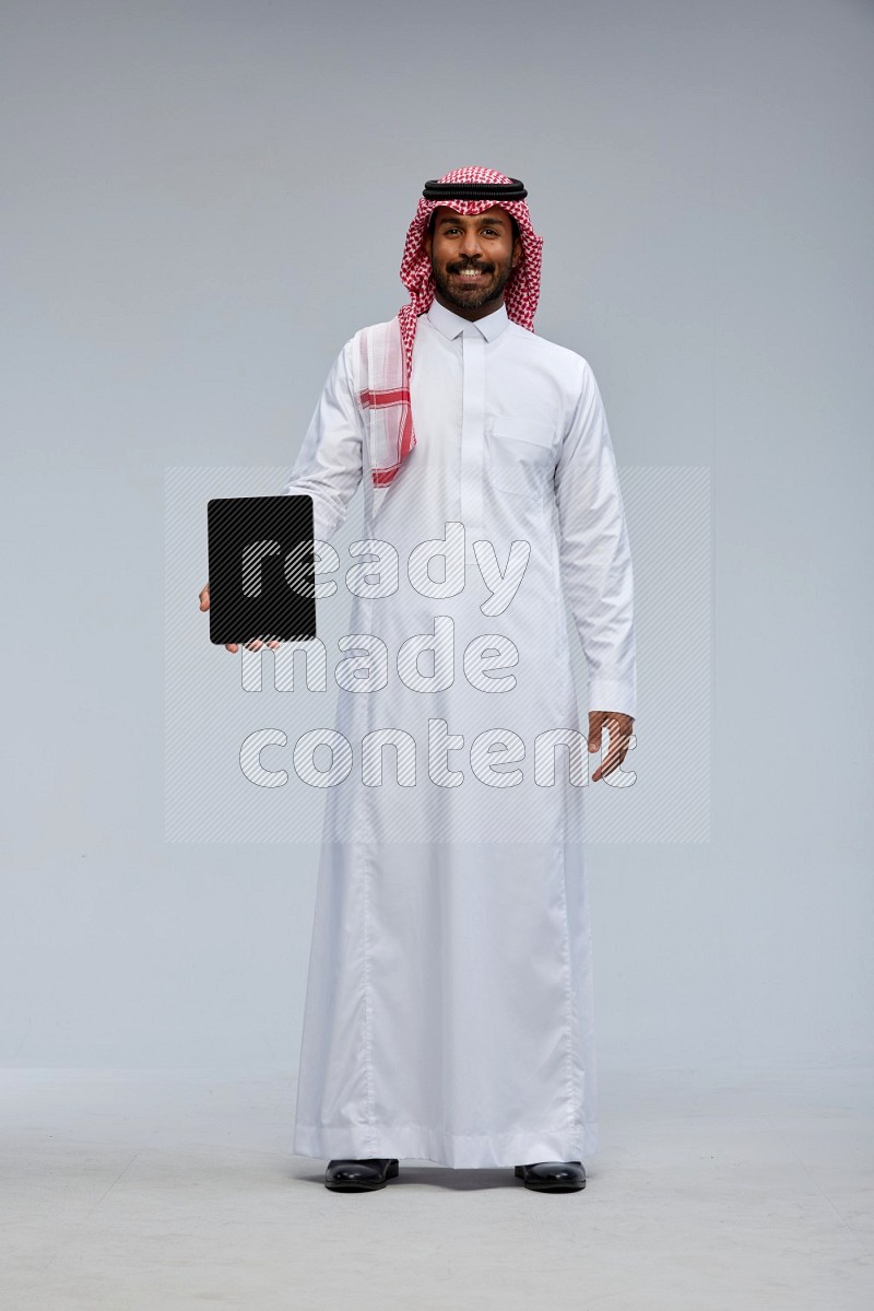 Saudi man Wearing Thob and shomag standing showing tablet to camera on Gray background